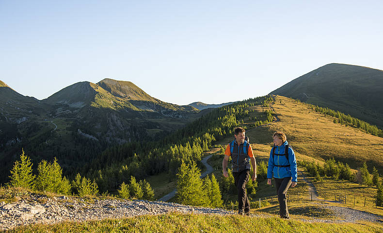  ALT-Text: Familie mit Kindern bei einer Wanderung in den Nockbergen, die glücklich an einem Gipfelkreuz steht und die Aussicht genießt. Bild 2: Wandern in den sanften Nockbergen Bildbeschreibung: Zwei Wanderer, ein Mann und eine Frau, gehen auf einem schmalen Pfad entlang eines grasbewachsenen Bergrückens in den Nockbergen. Der Mann trägt einen orangefarbenen Rucksack und eine graue Weste, die Frau eine blaue Jacke. Sie unterhalten sich und genießen die Bewegung in der Natur. Die Landschaft ist geprägt von sanften, grünen Hügeln und im Hintergrund sind weitere Gipfel der Nockberge zu sehen. Die Sonne scheint und taucht die Szene in ein warmes, goldenes Licht, das die friedliche und idyllische Atmosphäre des Wanderns unterstreicht. ALT-Text: Zwei Wanderer auf einem Wanderweg in den sanften, grasbewachsenen Nockbergen bei sonnigem Wetter.