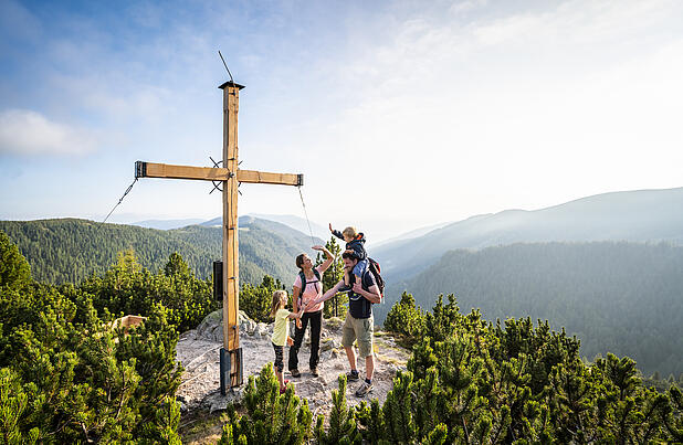 Familie mit Kindern bei einer Wanderung in den Nockbergen, die glücklich an einem Gipfelkreuz steht und die Aussicht genießt.